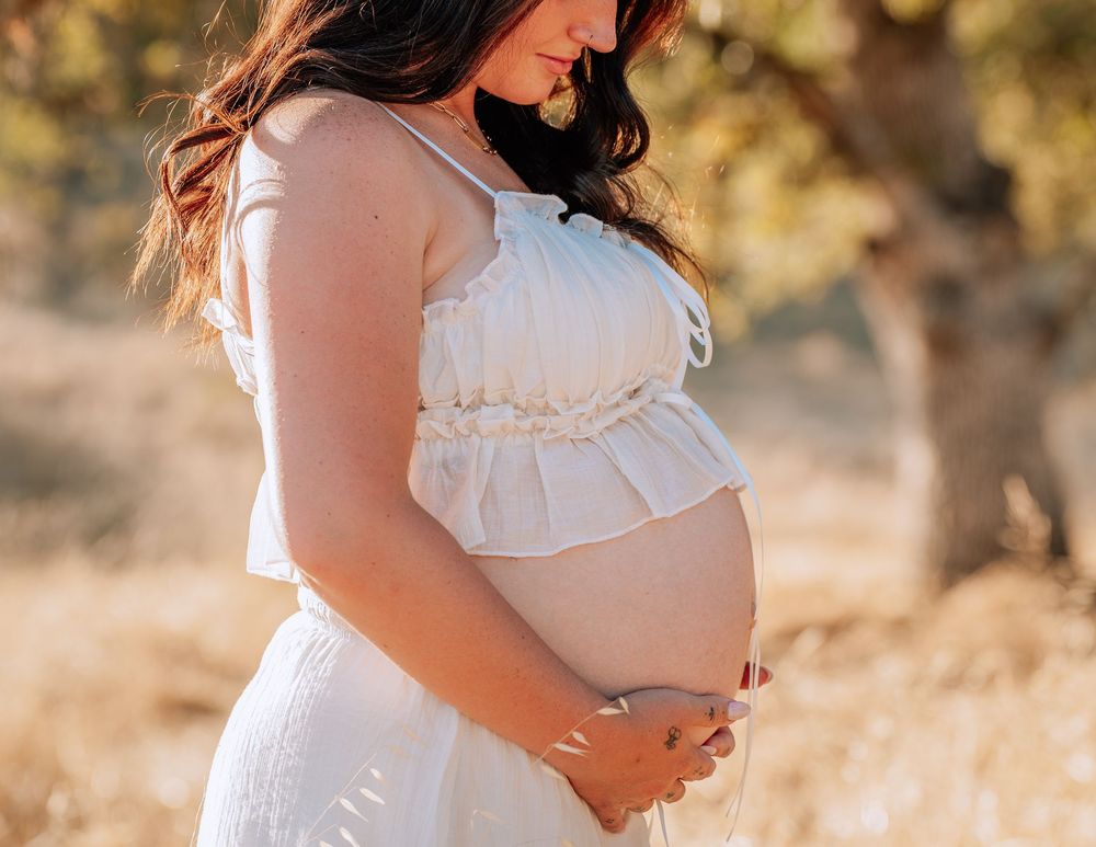 Pregnant woman in white dress cradling baby bump outdoors in golden sunlight during maternity photo session.