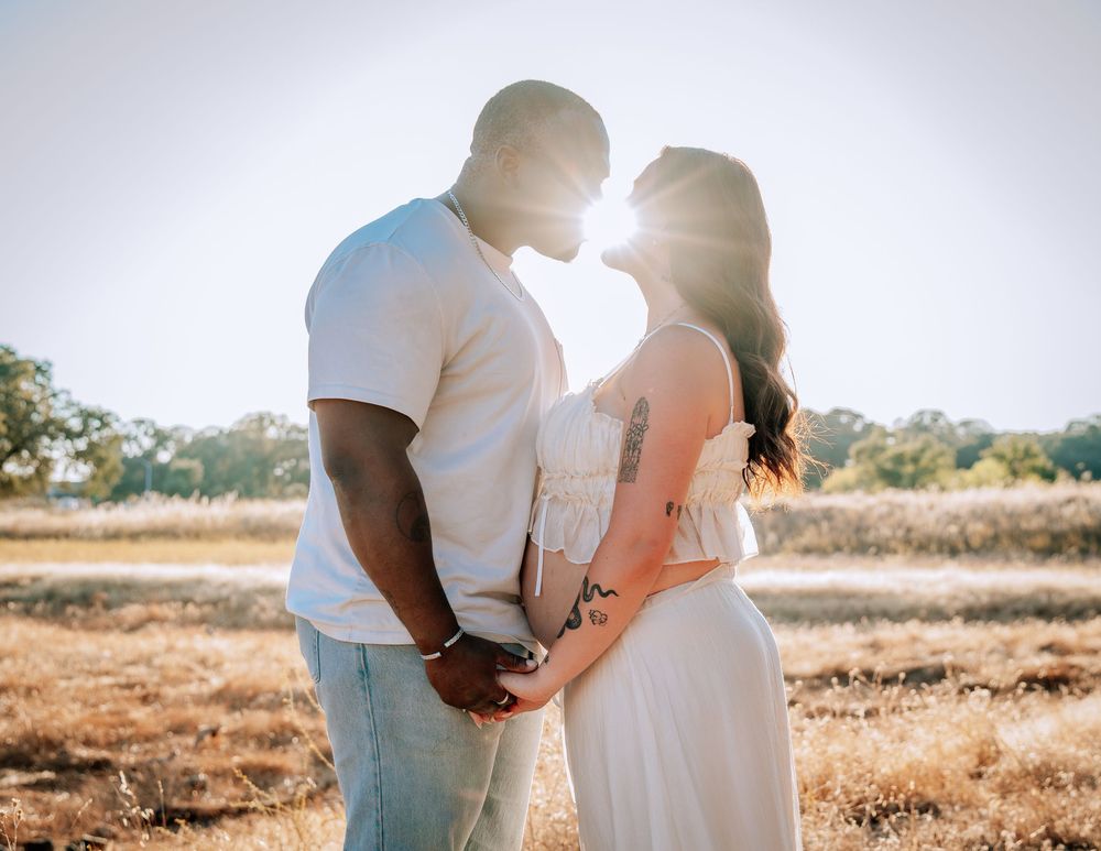 Couple sharing a romantic moment in a sunlit field, backlit by golden sun, man in white shirt and woman in white dress.