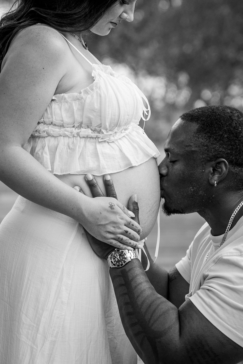 Black and white maternity photo of a man kissing pregnant woman's belly outdoors.
