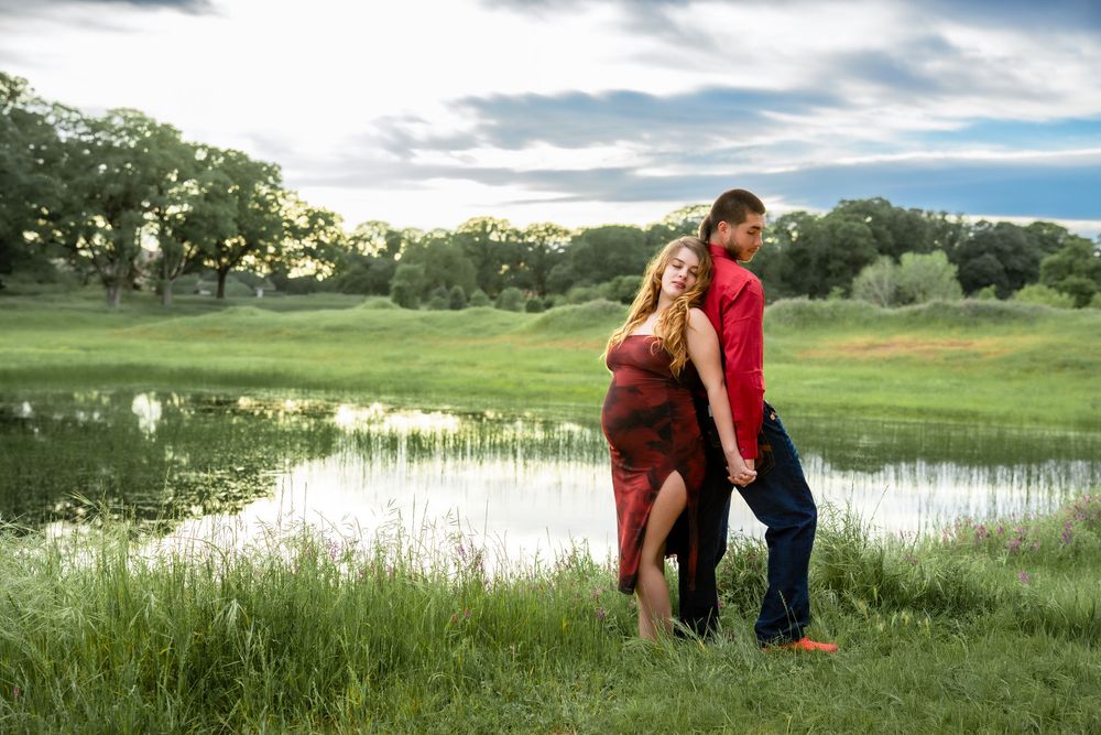 A pregnant woman in a red dress and a man embrace in a grassy field near a pond.