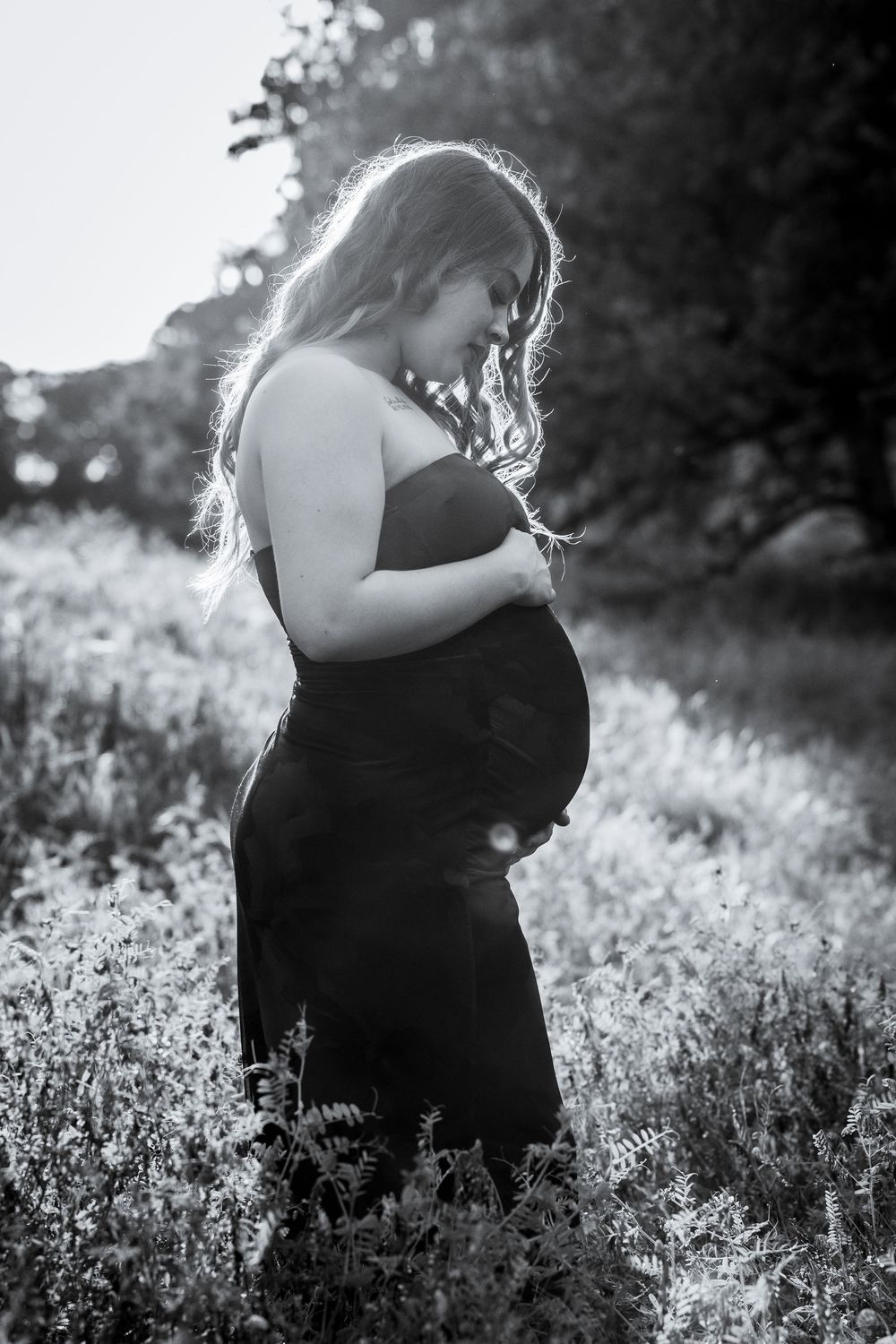 A pregnant woman stands in a field, her silhouette visible against the natural landscape.
