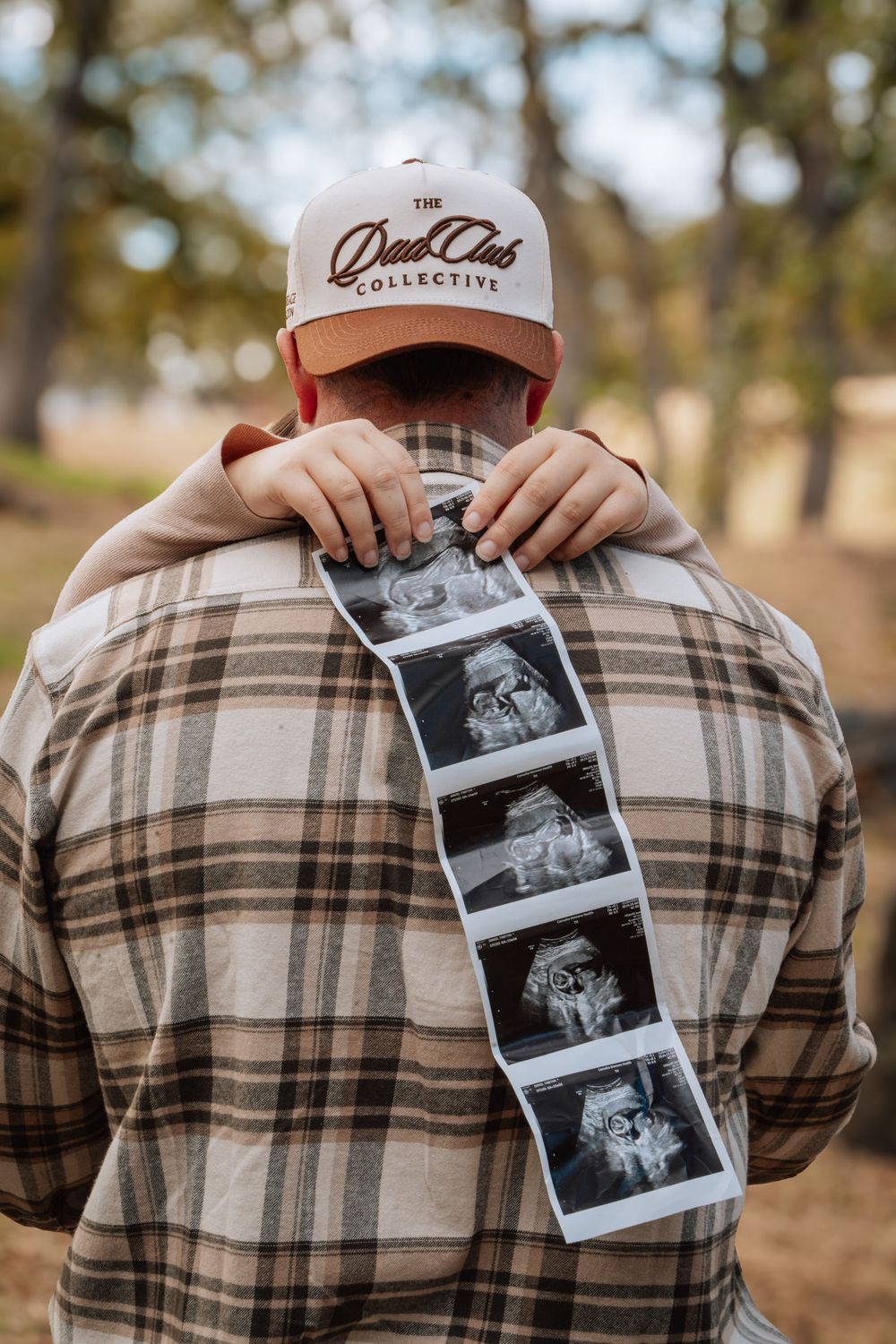 Someone wearing a plaid flannel shirt holds black and white ultrasound photos in a natural outdoor setting.