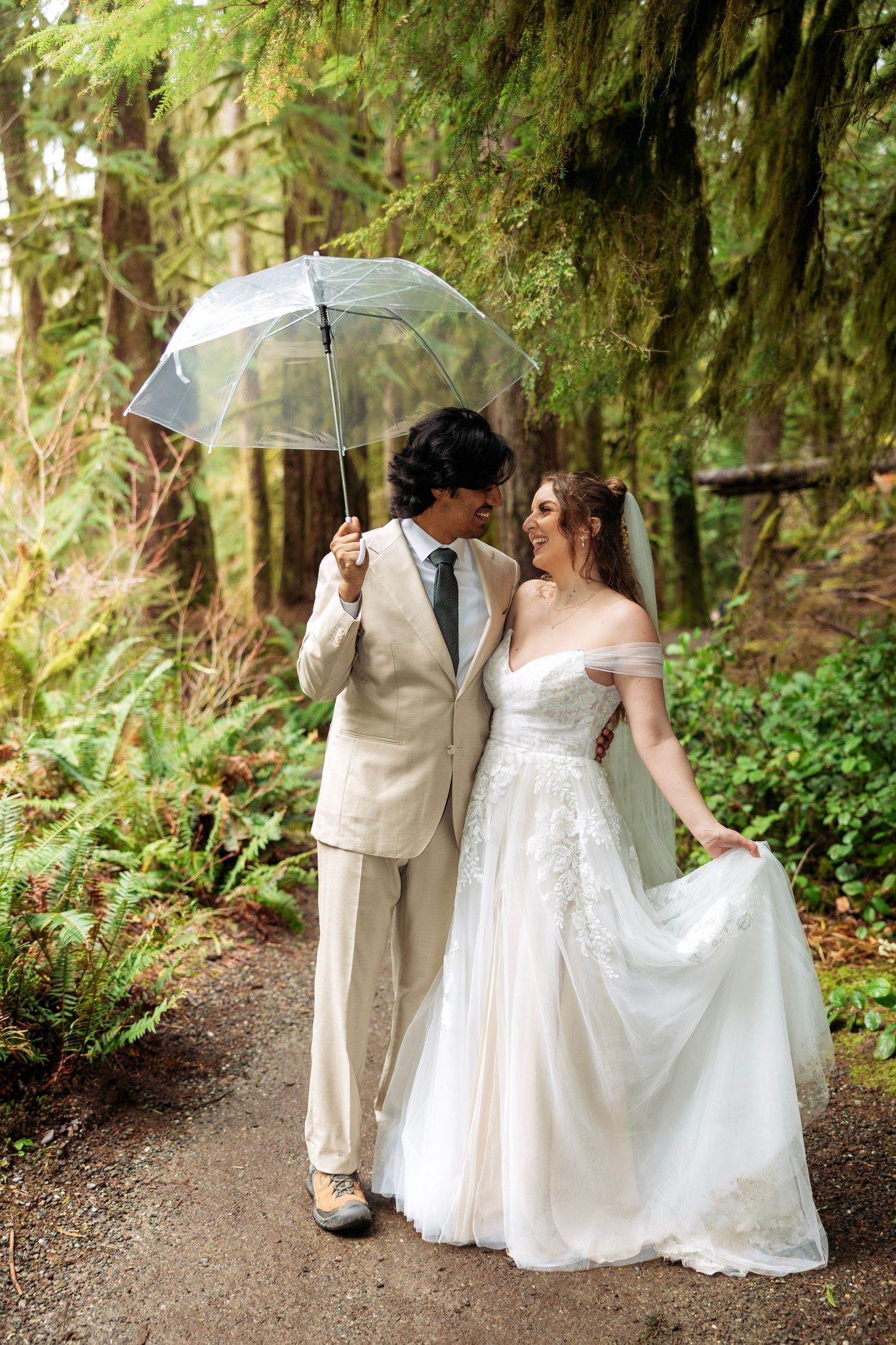 Romantic wedding couple walking together on a forest path holding a white umbrella in lush green surroundings.