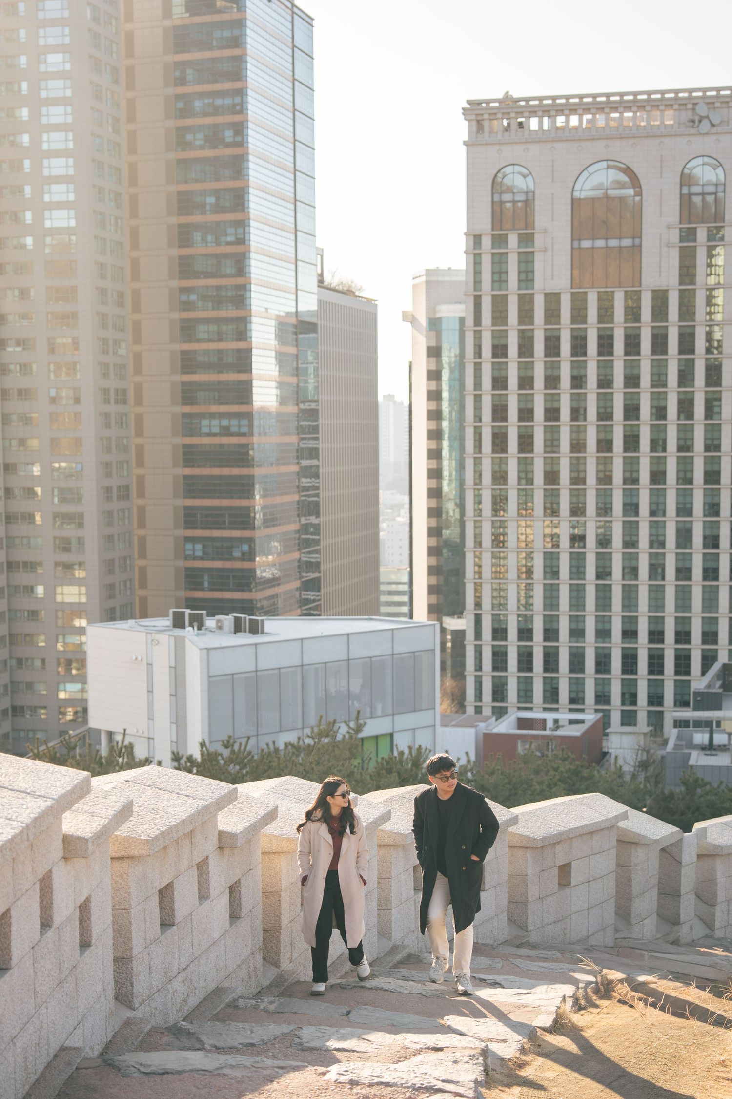 Couple walking along a rooftop terrace with urban high-rise buildings in the background during sunset.
