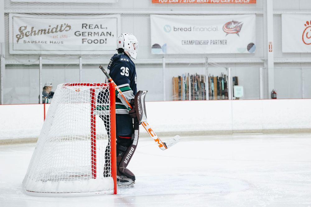 Ice hockey goalie in dark jersey positioned in front of red and white goal net.