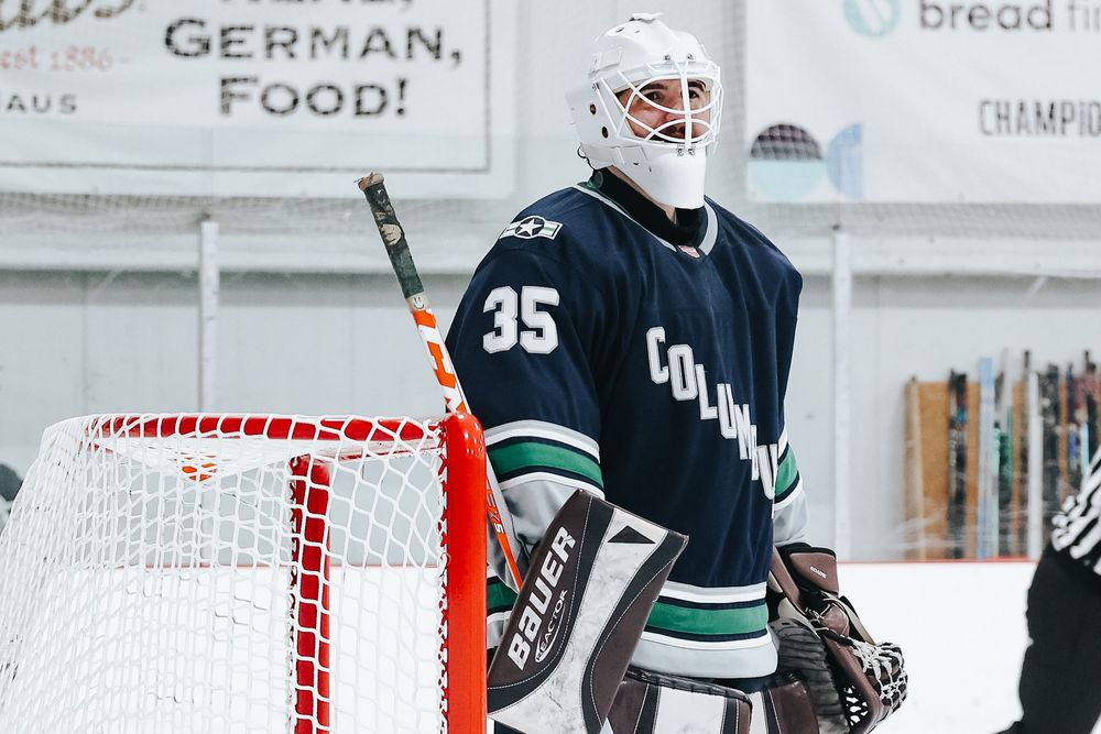 Hockey players in navy and green uniforms positioned near goal during game.