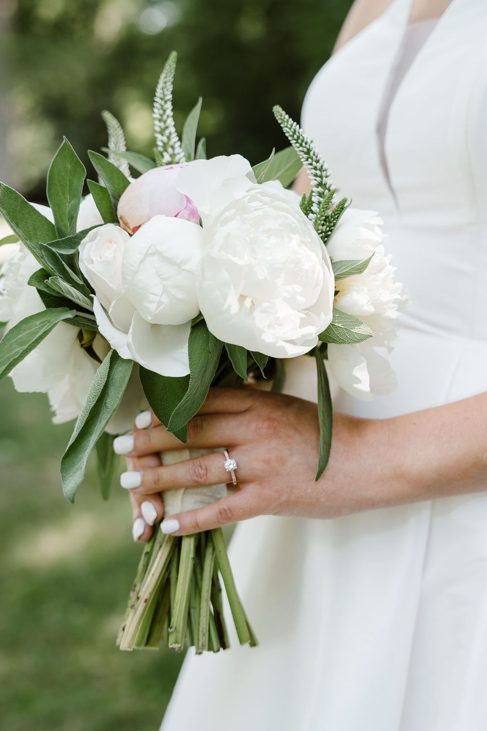 A delicate white peony and greenery bridal bouquet held by hands with an engagement ring.