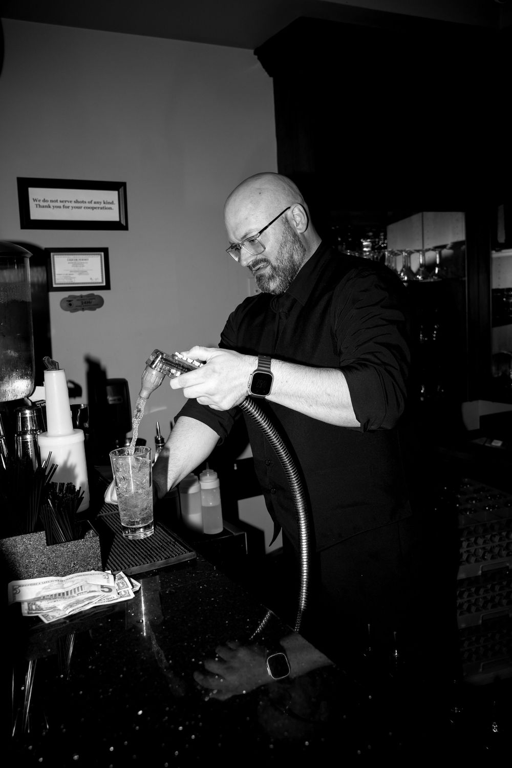A bartender in black attire pours a drink at a dark bar counter in black and white photography.