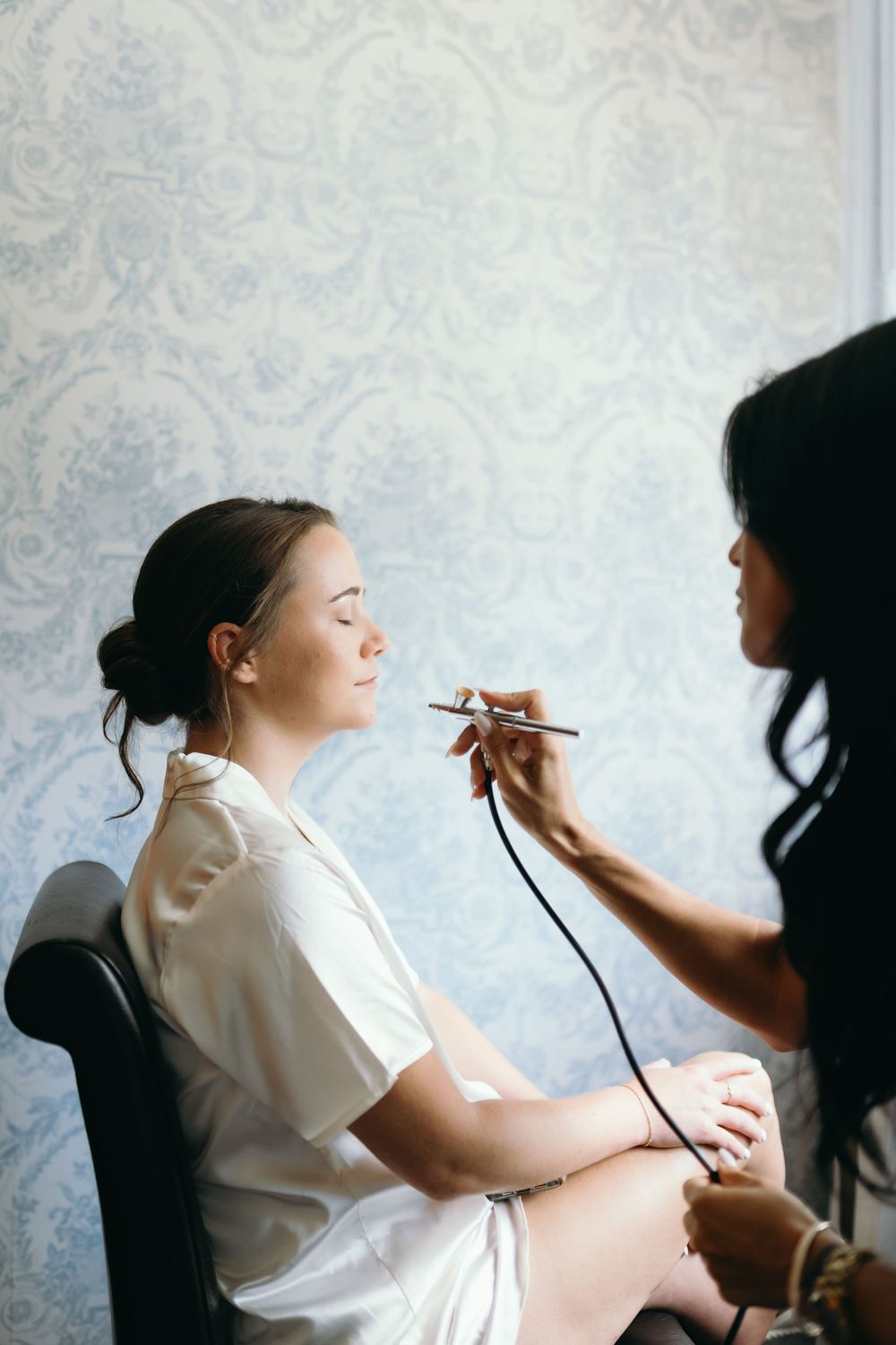 Someone getting their makeup professionally applied while sitting in a white chair against a patterned wallpaper background.
