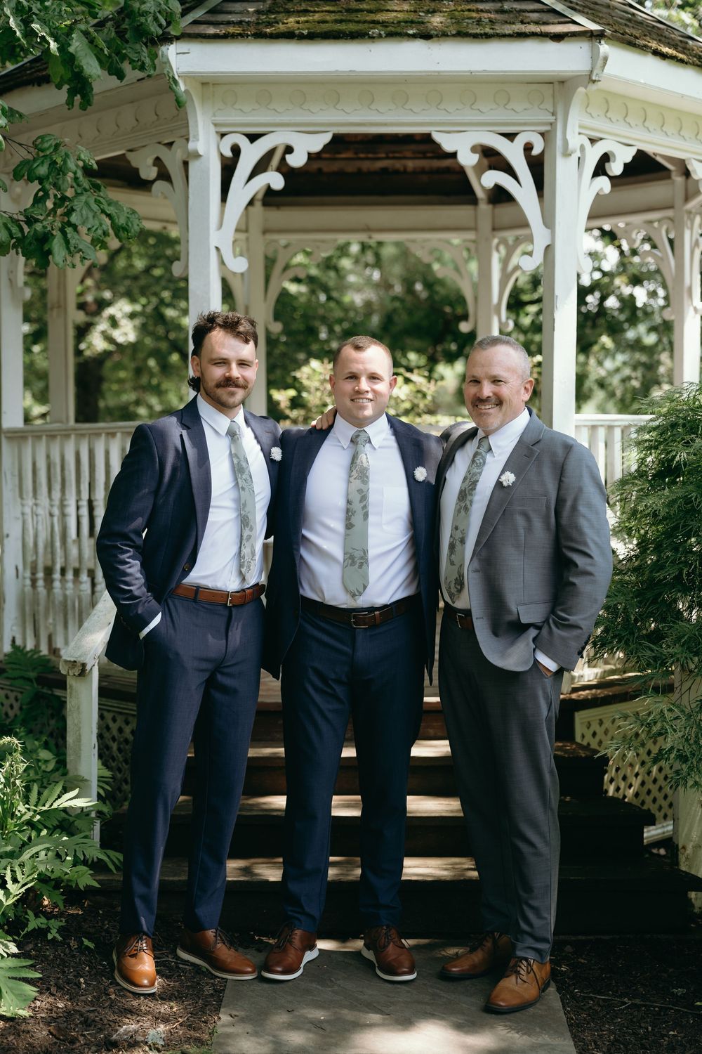 Groomsmen in matching navy suits and brown shoes pose together under a white garden gazebo.