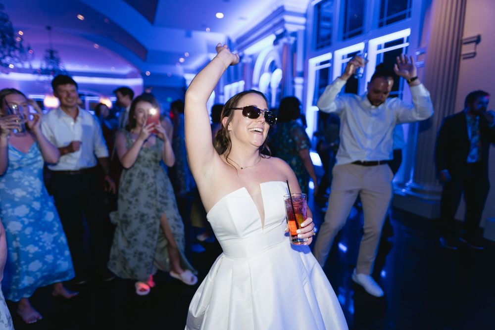 A person in a white wedding dress and sunglasses dances at a reception with blue lighting and other guests in the background.