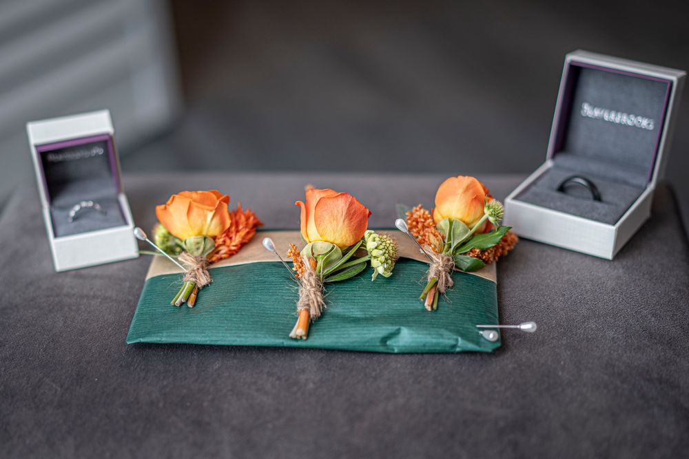 Wedding rings in boxes displayed alongside orange rose boutonnieres on green fabric.