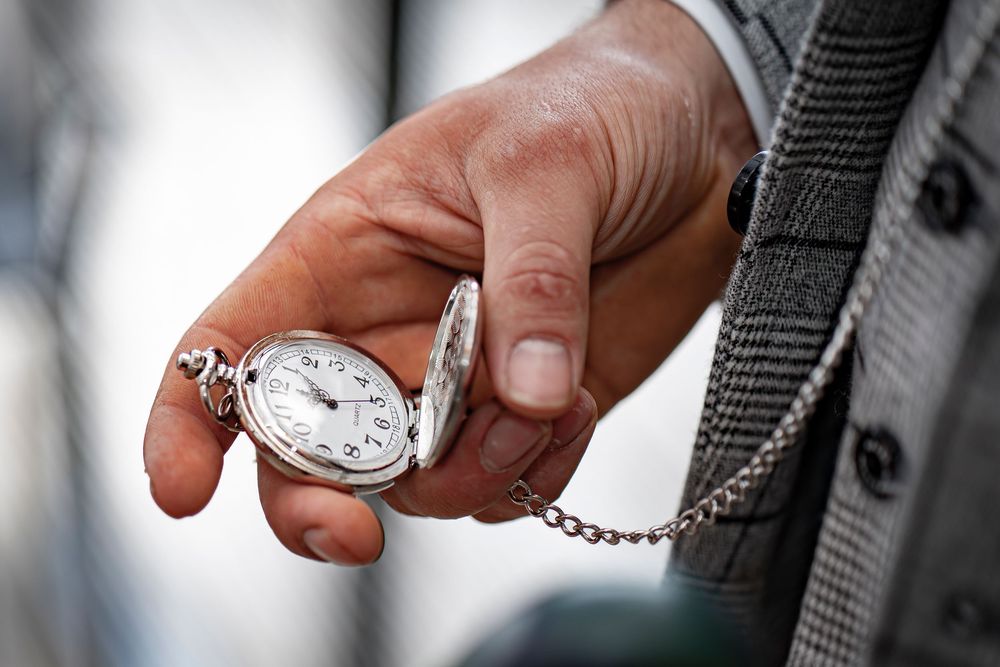 Hands holding a silver pocket watch on a chain against a blurred background.