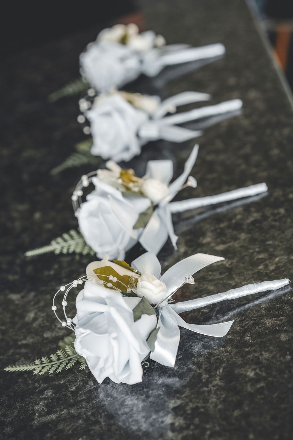 Row of white floral boutonnieres with ribbons arranged on dark stone surface.