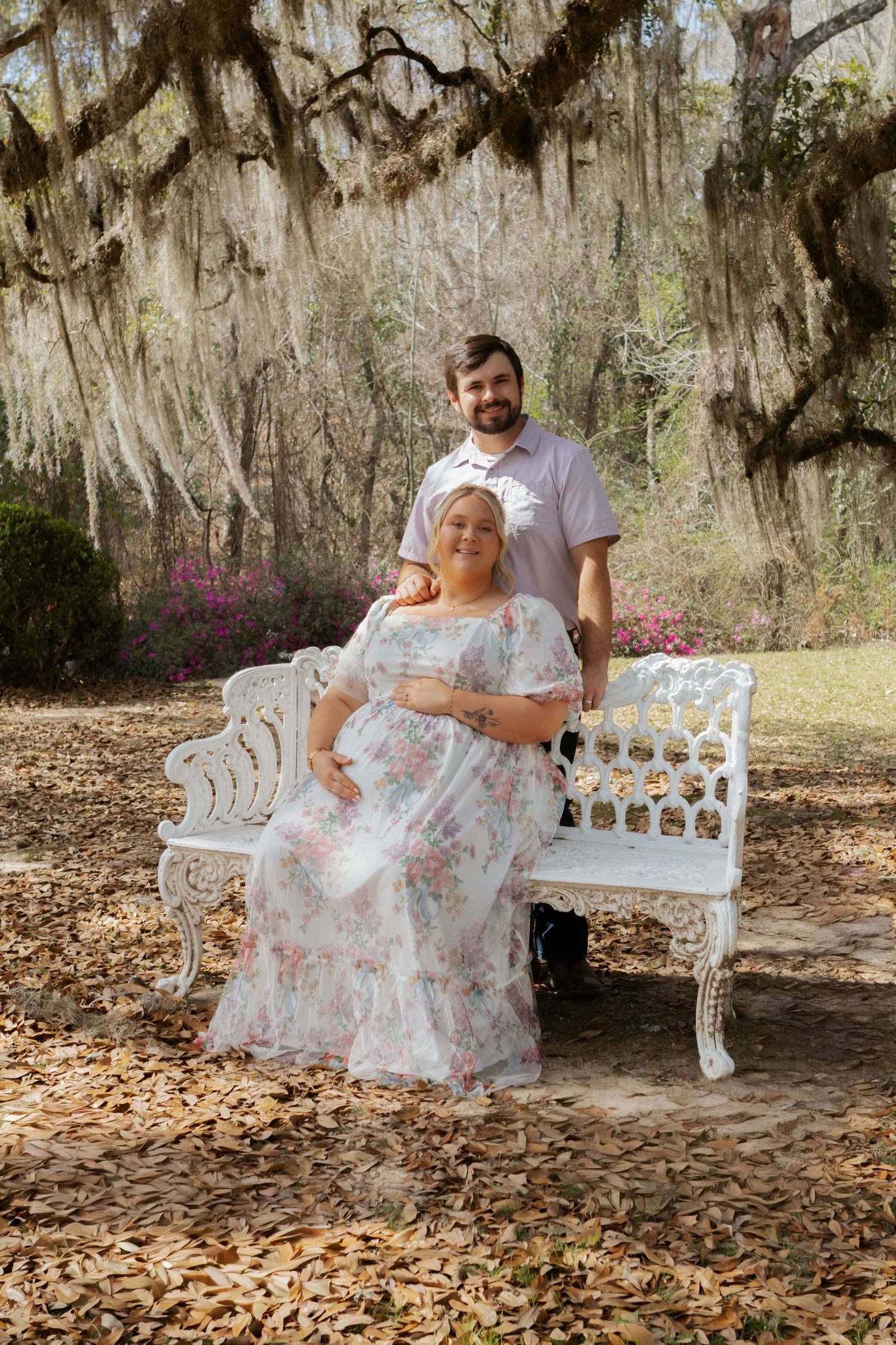 A couple shares a tender moment on a white garden bench under Spanish moss-draped trees in a romantic outdoor setting.