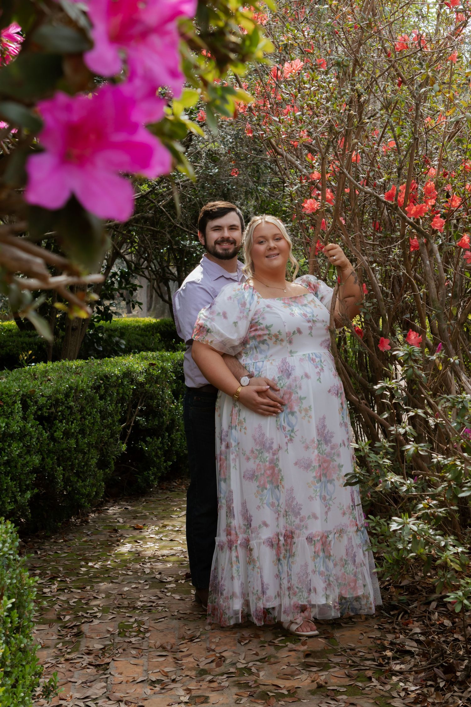 A couple poses together in a garden with pink flowers and green foliage creating a romantic outdoor setting.