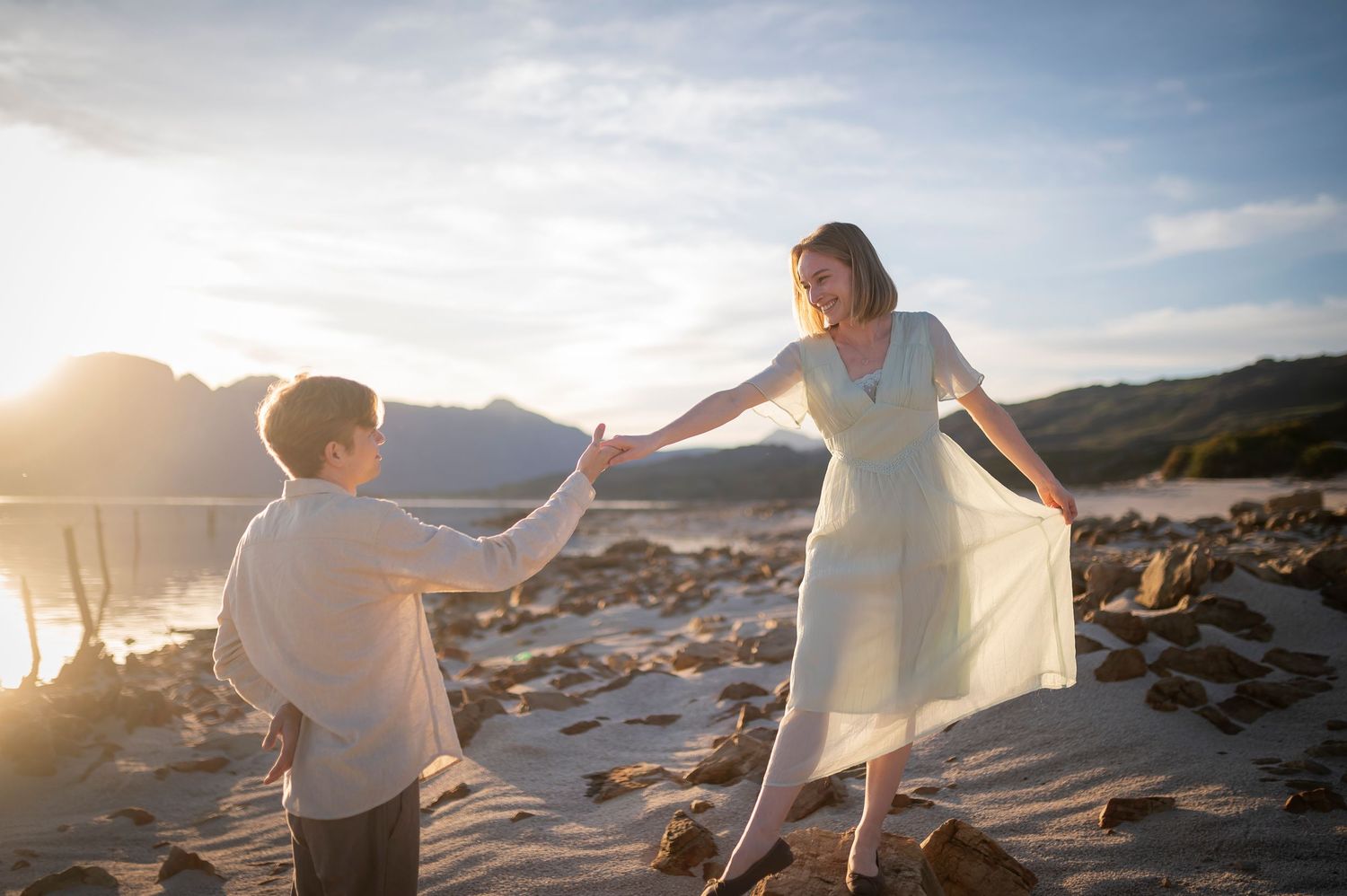 A woman in a white dress holds hands with a boy on a sunny beach with mountains in the background.