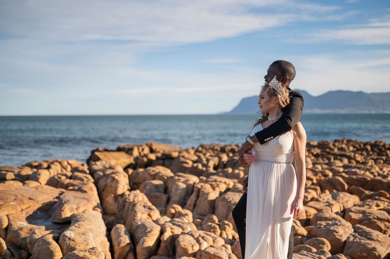 Pregnant woman in white dress standing on rocky coastline with ocean and mountain views.