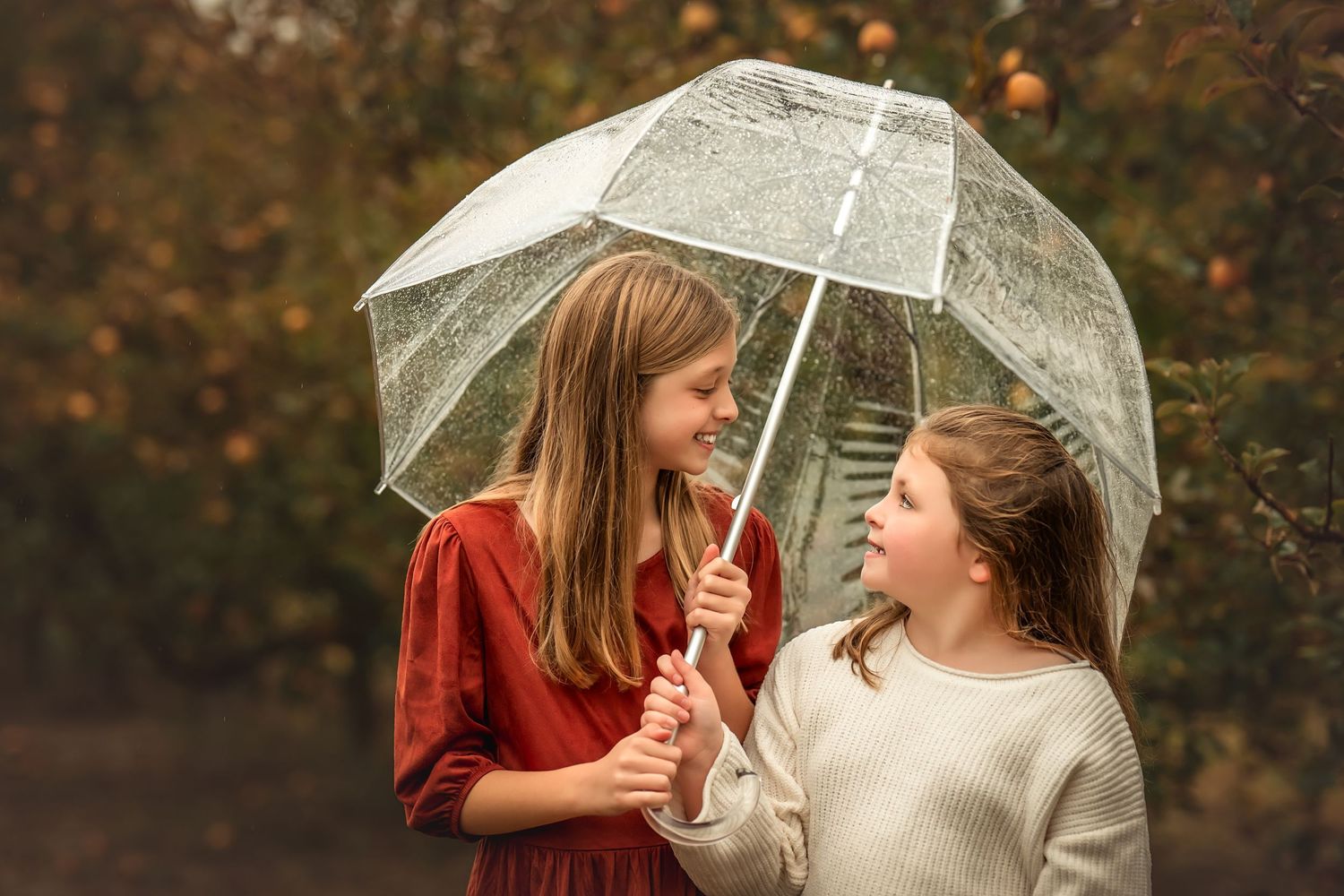 Two people in matching mint green dresses hold a clear umbrella while standing in an autumn field.