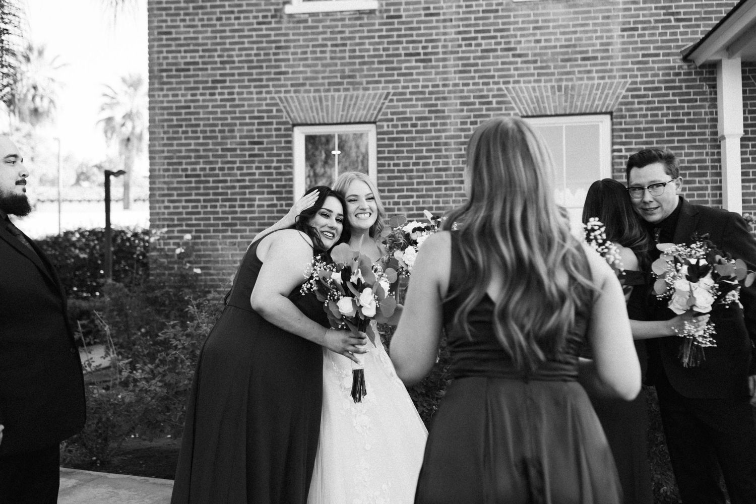 A black and white candid wedding party moment outside a brick building with people holding bouquets. Micro wedding, intentional celebrations