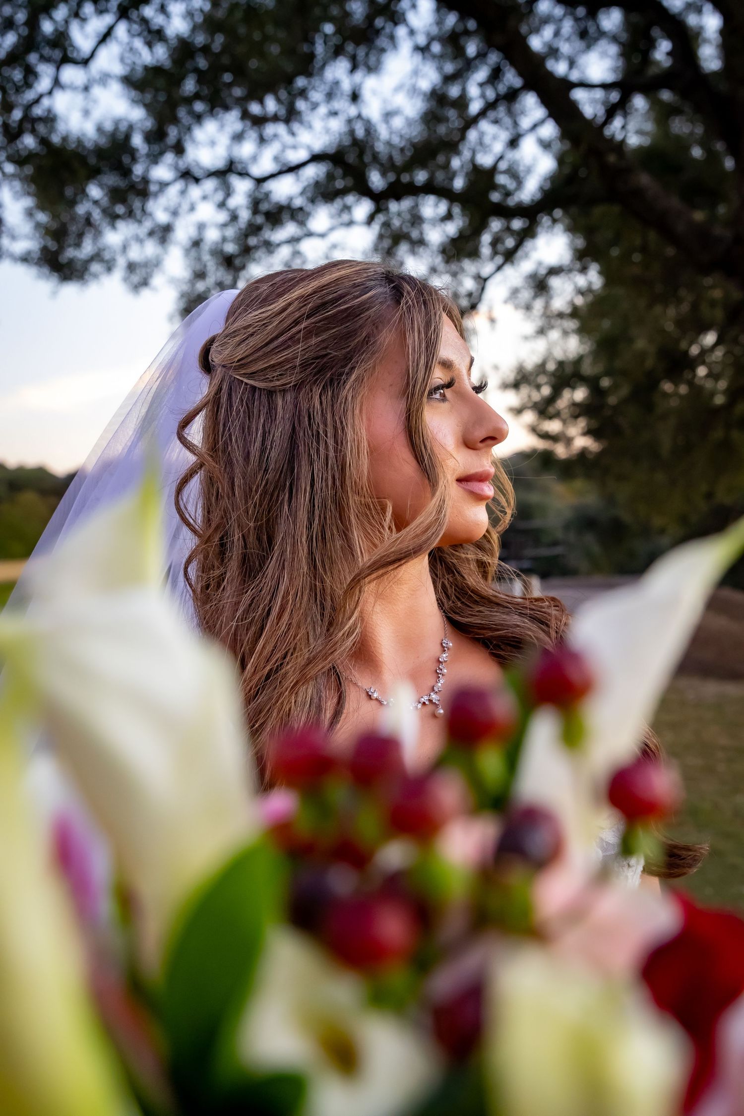 A couple in wedding attire sits together on a grassy hill at sunset, with a long flowing veil trailing behind them.