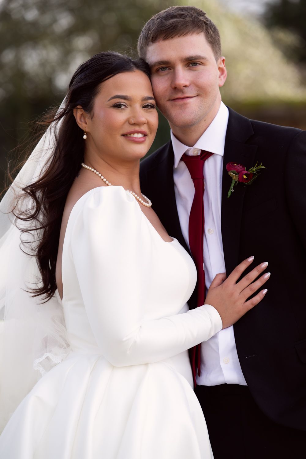 A couple in wedding attire poses together outdoors, with the bride in a white dress and groom wearing a red tie.
