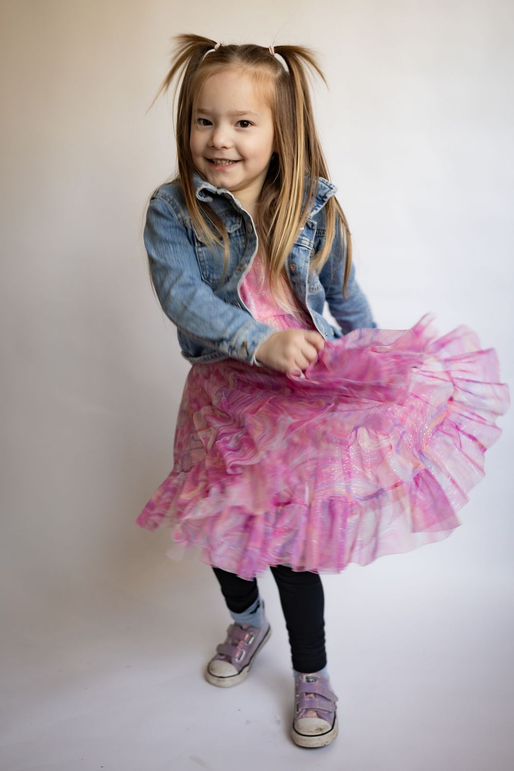A cheerful young girl twirls in a pink tutu dress and denim jacket with pigtails.