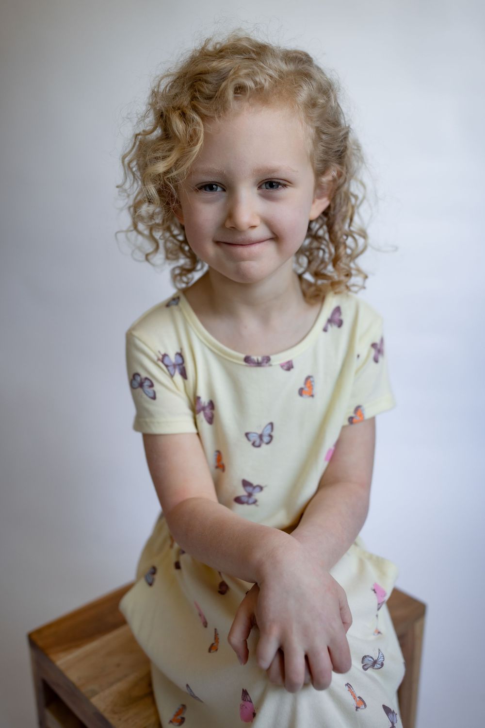 A curly-haired girl in a yellow butterfly dress sits smiling on a wooden stool.