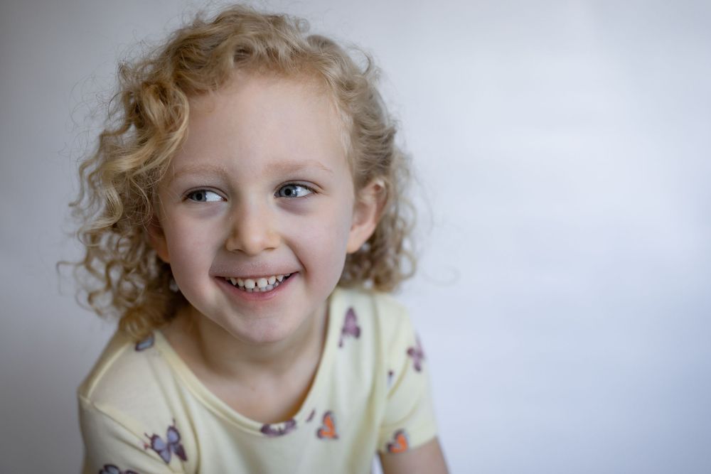 A smiling young girl with curly blonde hair wearing a yellow butterfly print shirt.