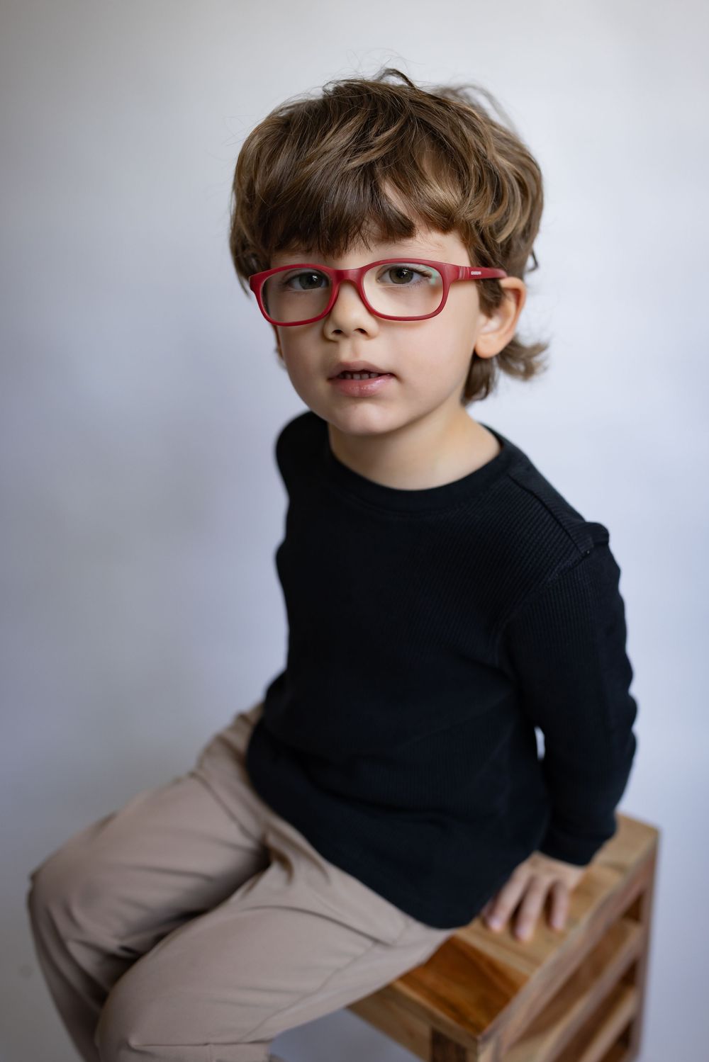 A young boy wearing red glasses and a black sweater sits on a wooden crate.