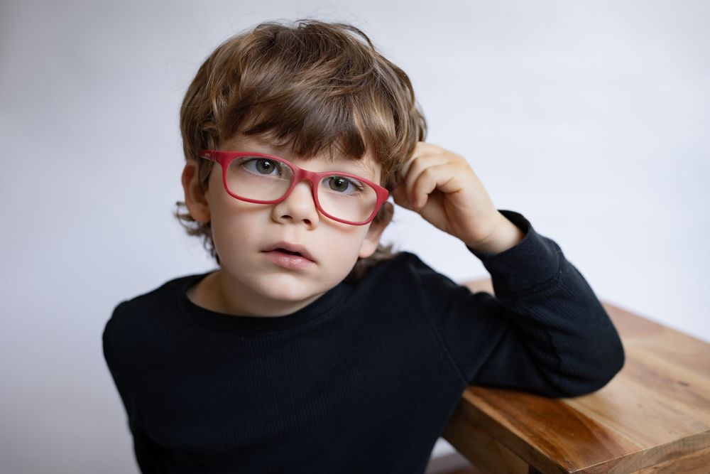 A young boy with red glasses rests his chin on a wooden surface thoughtfully.