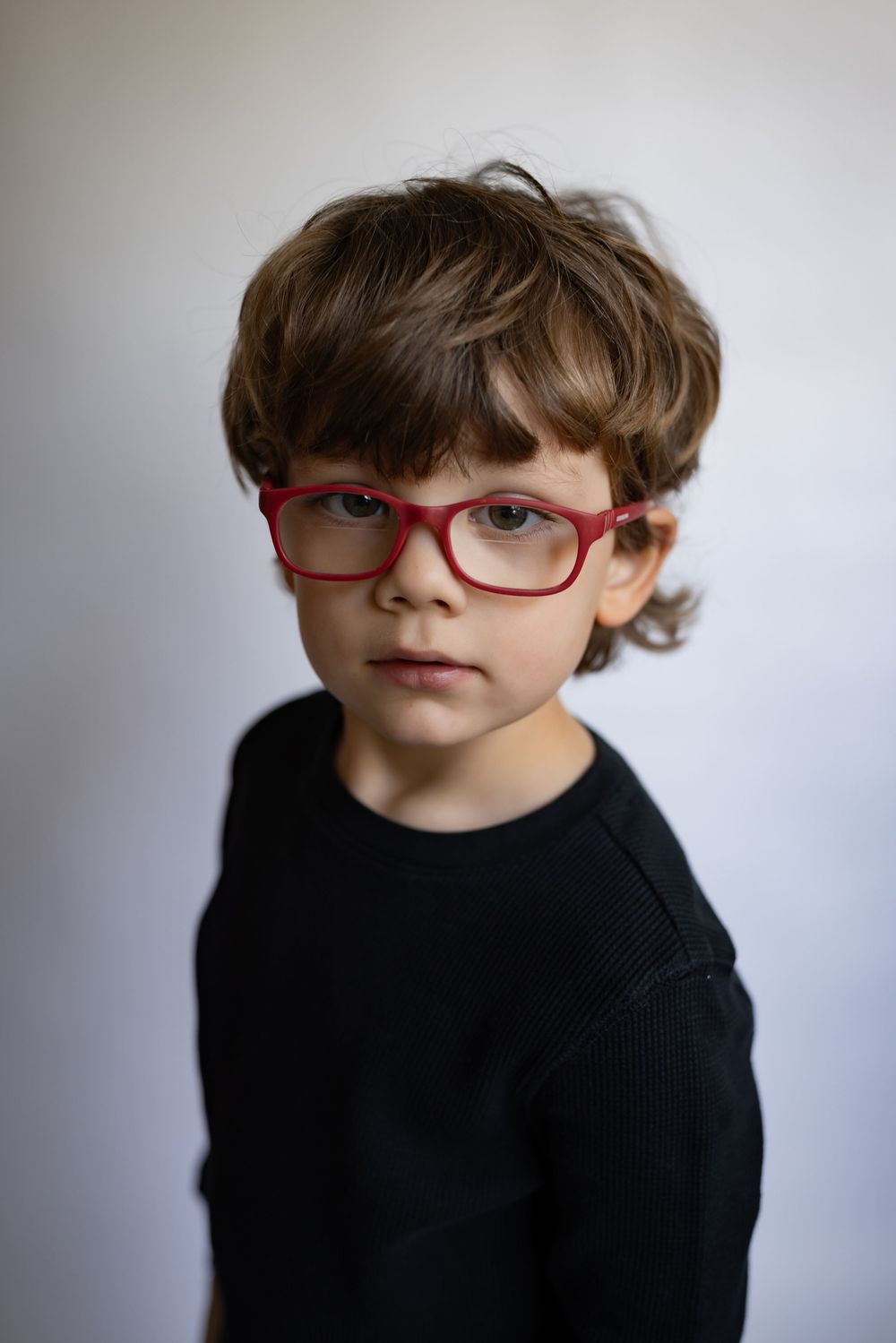 A serious young boy with red rectangular glasses wearing a black sweater poses for portrait.