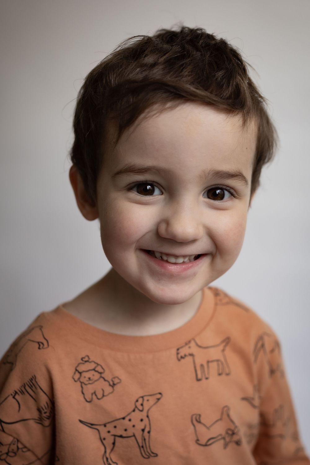 A smiling young boy in an orange dog-print shirt poses for a cheerful portrait.