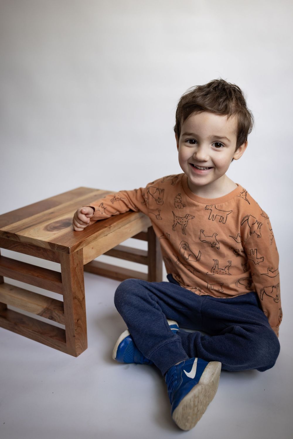 A happy young boy in an orange animal-print shirt sits cross-legged beside a wooden crate.