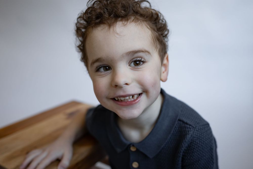 Smiling young boy with curly hair leaning on a wooden bench against a white background.