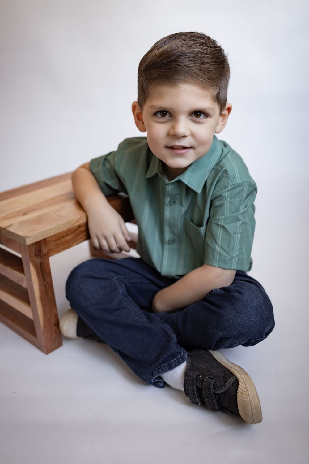 Young boy in green shirt and jeans sitting cross-legged on floor beside wooden crate, studio portrait.