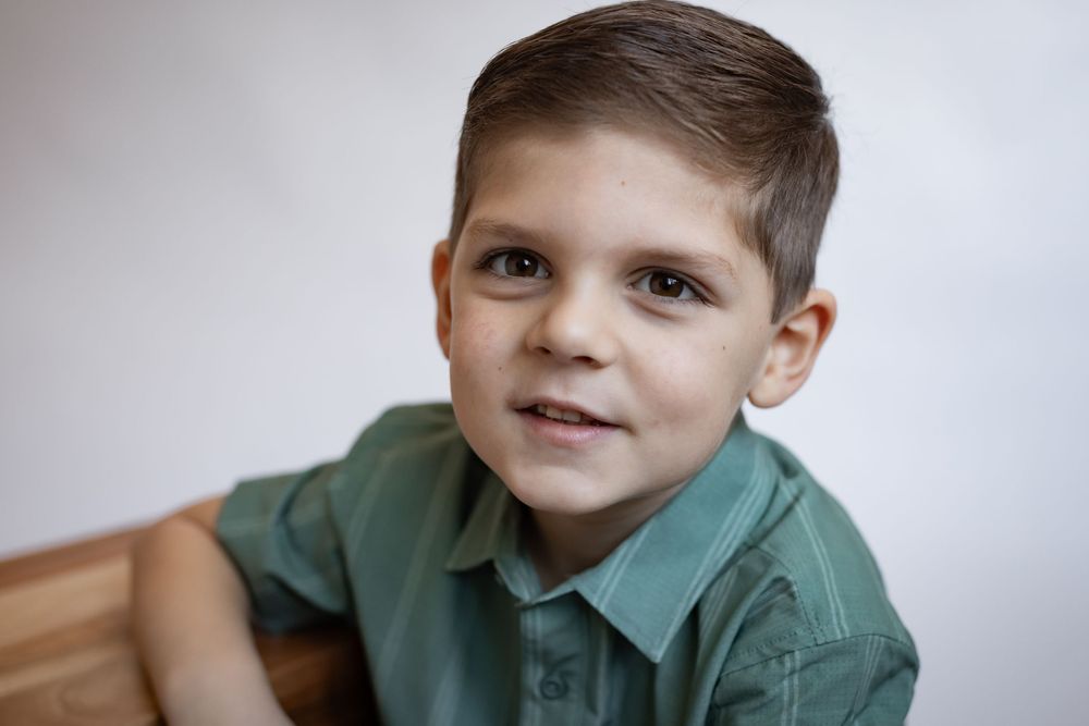 A young boy with brown hair smiles warmly at the camera, wearing a green polo shirt.