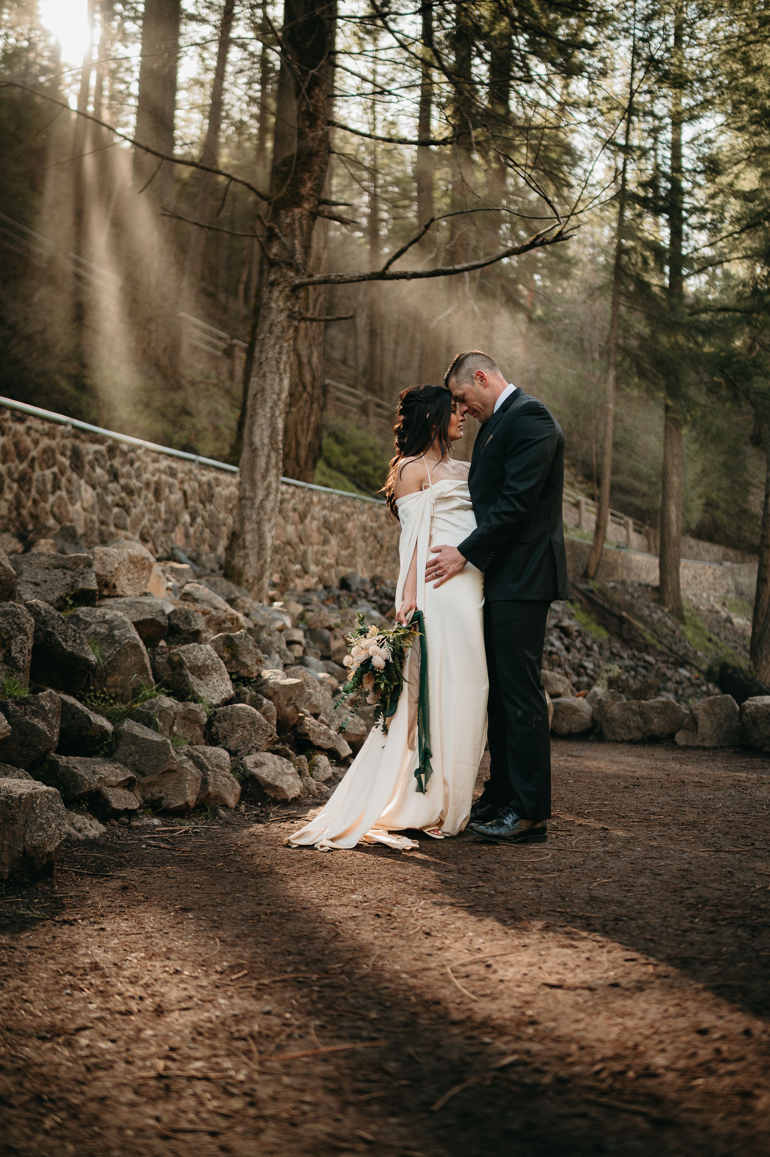 Romantic wedding couple embraces in a misty forest with sunbeams streaming through tall evergreen trees.