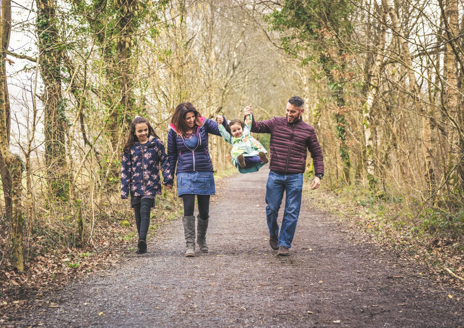 A fun family photo shoot. Mum, Dad and 2 daughters walking down a woodland path, with Mum and Dad swinging the younger daughter in the air.