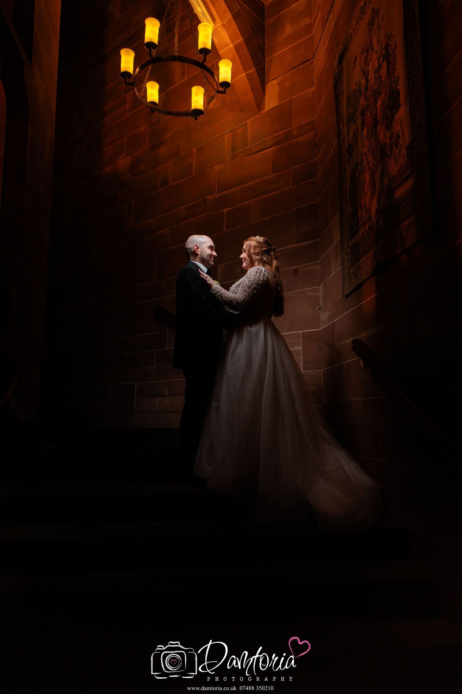 A couple in formal attire embraces in a dimly lit stone corridor with glowing yellow lanterns casting romantic shadows.