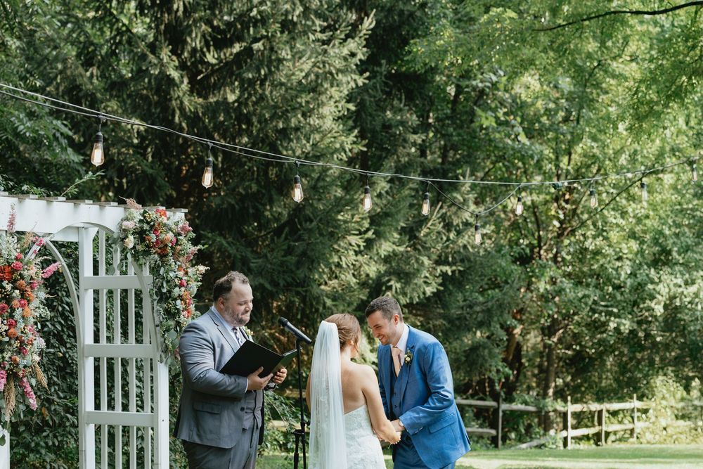 Couple shares intimate moments during outdoor wedding ceremony under lush green trees and white arbor.