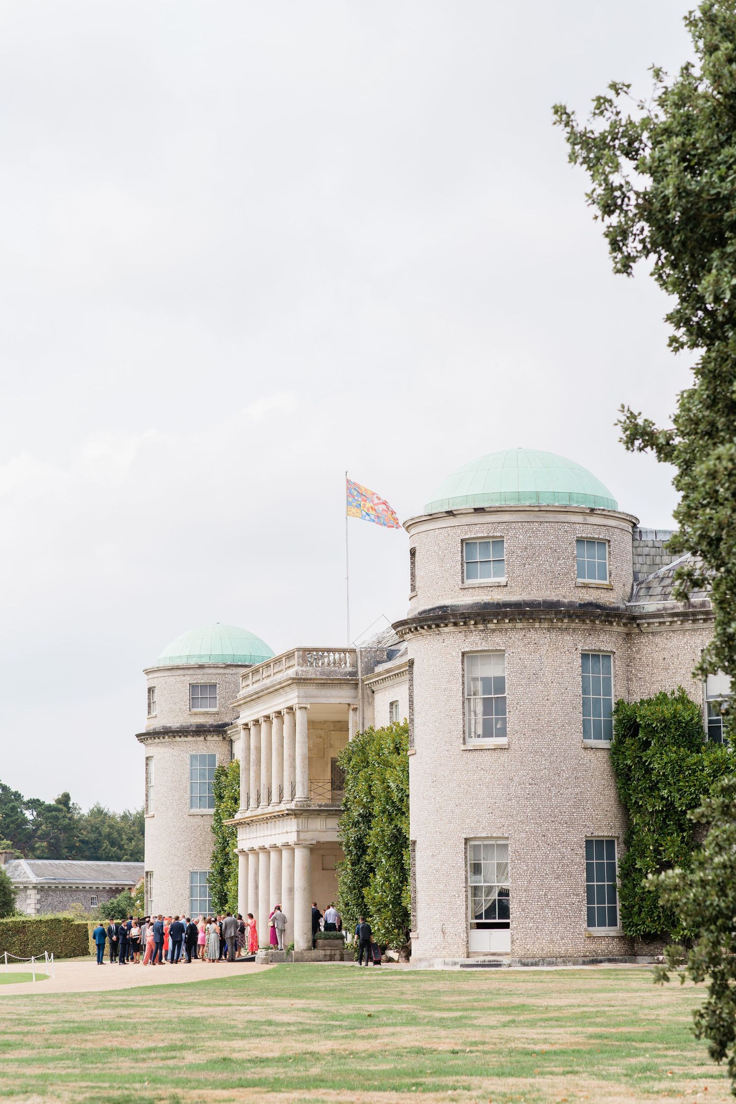 Elegant historic mansion with turquoise domed towers and neoclassical architecture stands on manicured lawn with visitors gathered.