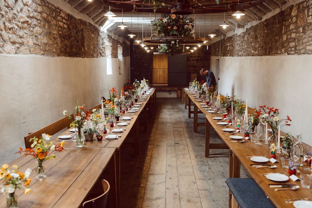 Long rustic dining tables set for an elegant dinner with floral centerpieces in a stone-walled cellar venue.
