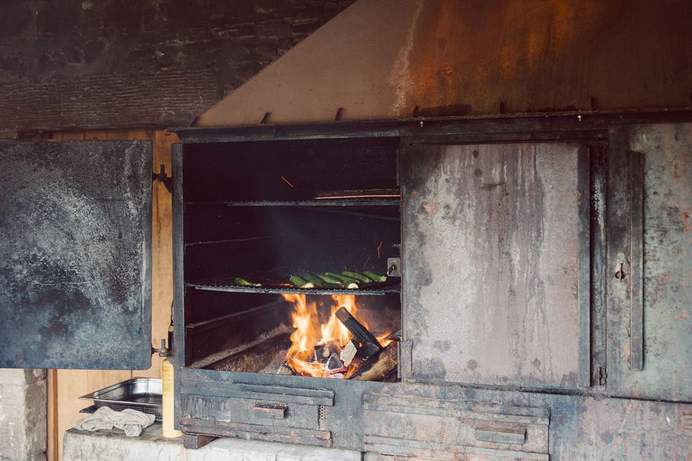 A rustic outdoor fireplace with burning logs and flames inside a weathered metal and concrete structure.