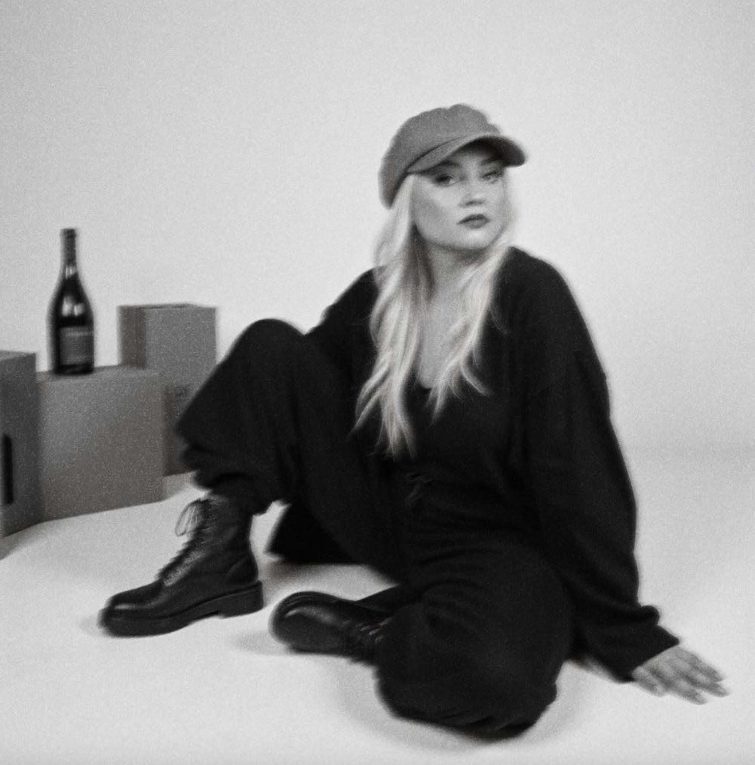 Black and white photo of blonde woman in black outfit and cap sitting on floor near wine bottle.