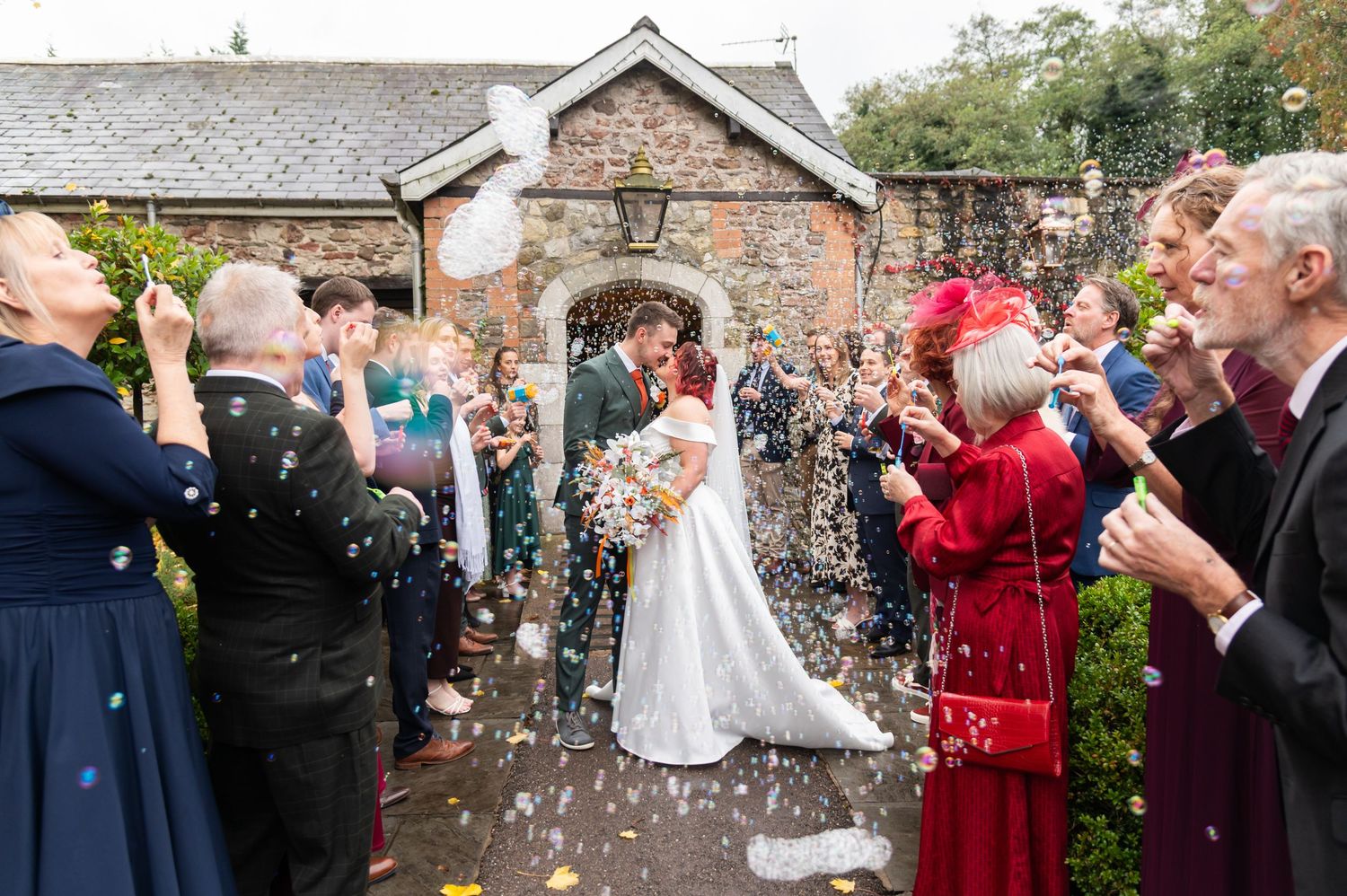 Wedding guests throw colorful confetti at a couple outside a rustic stone building during a celebration.