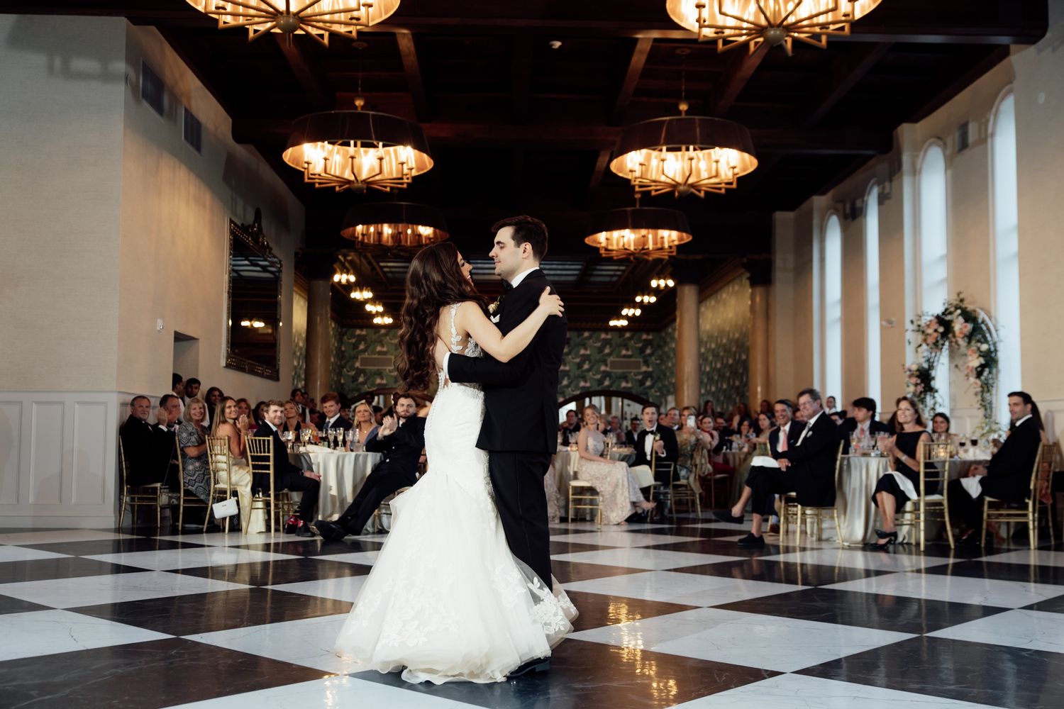 A couple shares their first dance on a black and white checkered ballroom floor under elegant crystal chandeliers.