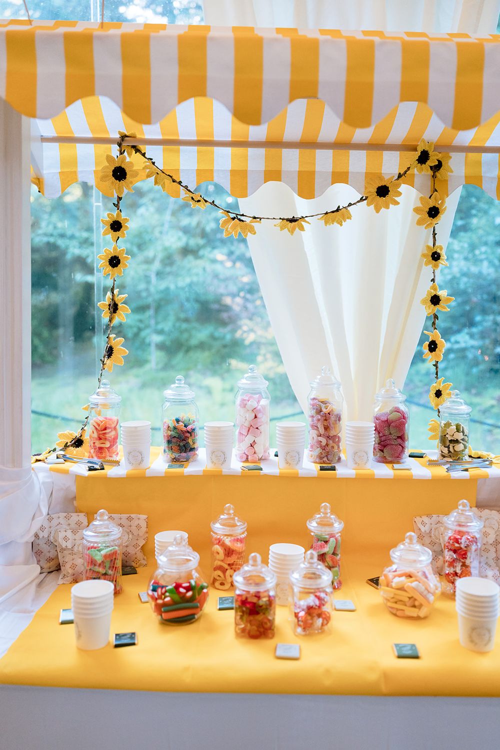 Glass candy jars with pink and white treats lined up on a yellow table with sunflower garland decorations above.