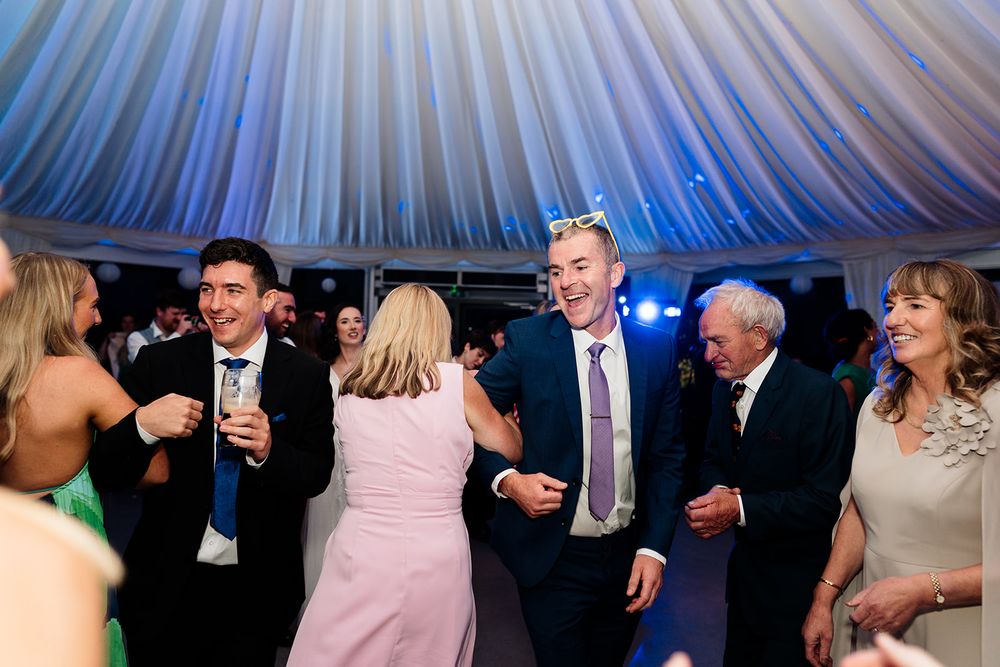 Wedding guests dancing under a blue and white tent with festive lighting and decorations.