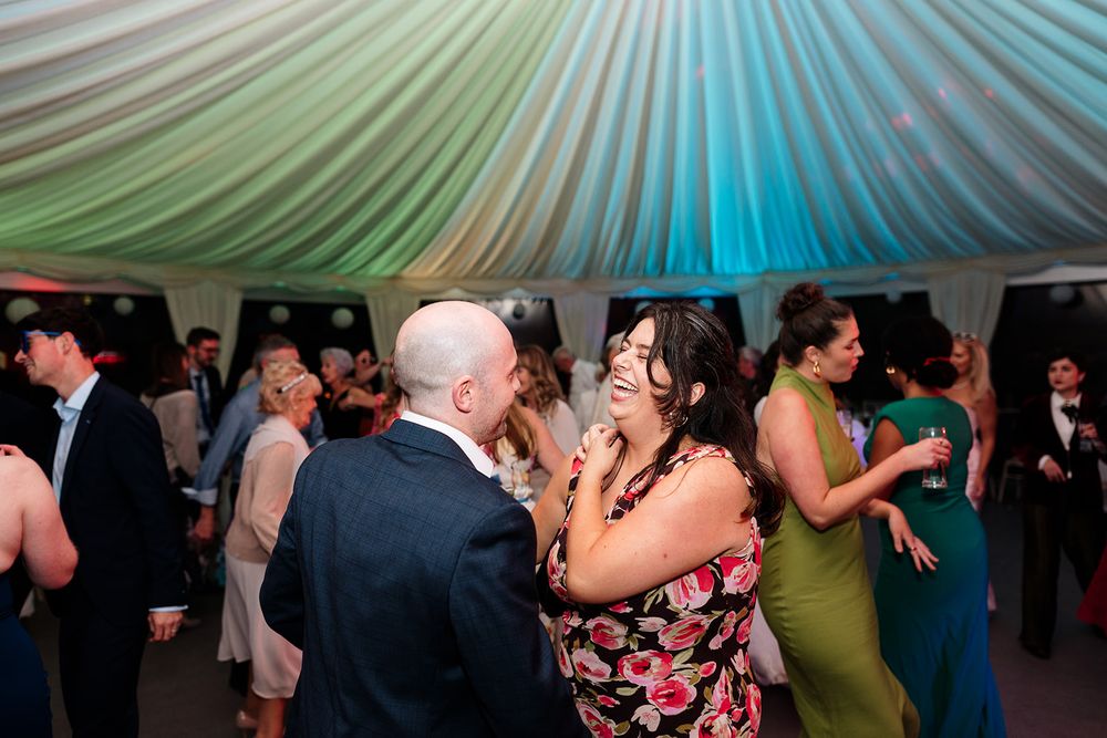 People dancing and celebrating at a wedding reception under a white tent with colorful lighting.